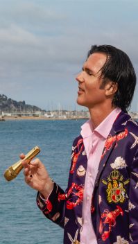 Der Künstler steht in rosa Hemd und buntem Jackett an einer Küste und hält ein goldenes Mikrofon wie eine Zigarre | Quelle: Landry