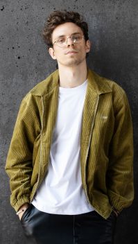 Tim Kamrad lehnt in einer grünen Jacke an einer grauen Betonwand. (Quelle: Bella Lieberberg)