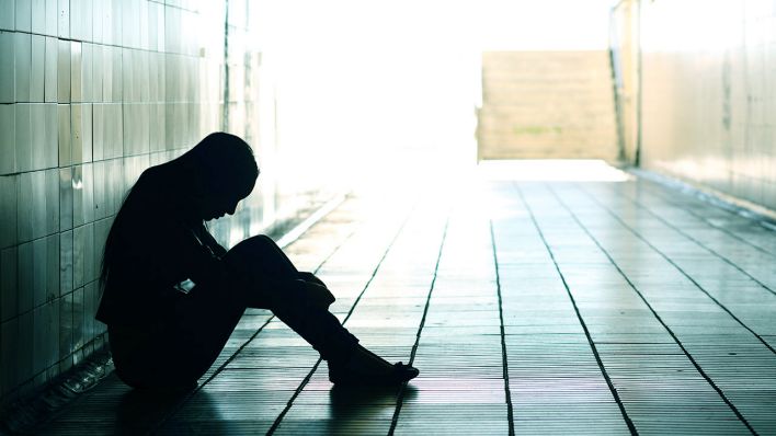 Symbolbild Mental Healt: Eine weiblich gelesene Person sitzt mit dem Rücken an der Wand auf dem Boden und umgreift ihre herangezogenen, aufgestellten Beine. Es ist nur ihre Silhouette zu erkennen. Symbolbild Mental Healt: Eine weiblich gelesene Person sitzt mit dem Rücken an der Wand auf dem Boden und umgreift ihre herangezogenen, aufgestellten Beine. Es ist nur ihre Silhouette zu erkennen.