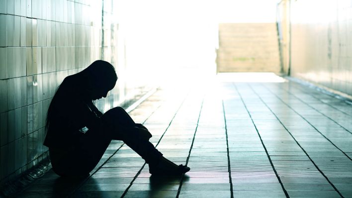 Symbolbild Mental Healt: Eine weiblich gelesene Person sitzt mit dem Rücken an der Wand auf dem Boden und umgreift ihre herangezogenen, aufgestellten Beine. Es ist nur ihre Silhouette zu erkennen.