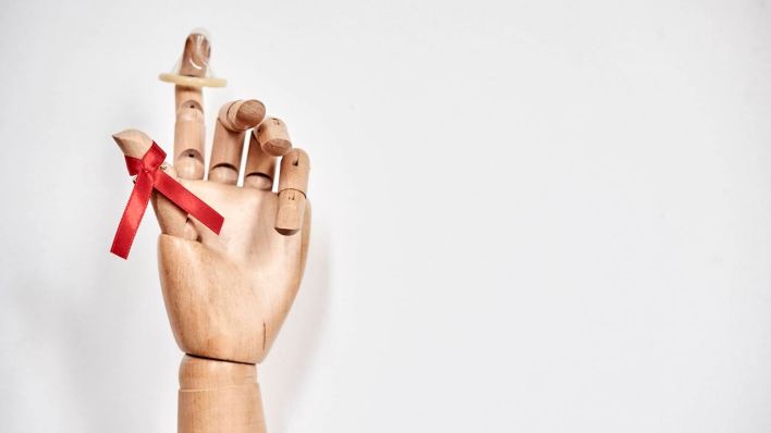 Auf den Fingern einer Holzhand stecken ein Kondom und eine rote Schleife. (Quelle: Picture Alliance/BSIP)