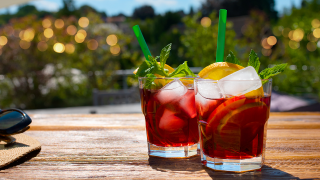 Zwei Gläser mit einem braunen Getränk, Eiswürfeln, jeweils einer Orangenscheibe und einem grünen Strohhalm stehen auf einem Holztisch. Im Hintergrund ist unscharf ein Garten mit Lichterketten zu erkennen. Die Sonne scheint. | Quelle: IMAGO / Panthermedia