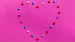 Ein Herz gelegt aus vielen kleinen Plastikherzen in den Farben blau, rot und weiß. Die Herzen liegen auf einem rosanen Untergrund. | Quelle: IMAGO / Wirestock