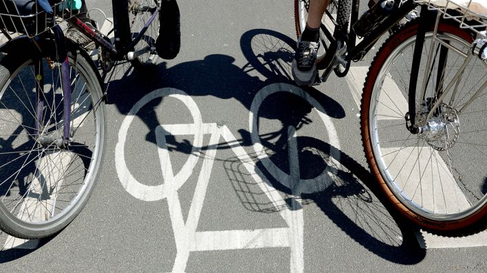 Links und rechts sind die Vorderreifen von zwei Fahrrädern zu sehen. Sie stehen auf einem Fahrradweg, auf dem ein Fahrradzeichen gemalt ist. | Quelle: IMAGO / Sabine Gudath