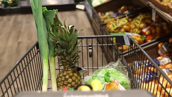 Eine Nahaufnahme von einem Einkaufswagen, in dem sich schon etwas Obst und Gemüse befindet. Im Hintergrund ist die Obst- und Gemüseabteilung eines Supermarktes zu erkennen. | Quelle: IMAGO / CHROMORANGE