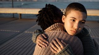 Zwei Personen umarmen sich auf einem Pier am Meer. Eine Person guckt traurig in die Ferne (Quelle: IMAGO / Westend61)