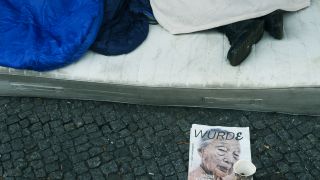 Ein Obdachloser Mann liegt auf einer Matratze und schläft. Es sind nur seine Füße zu sehen. Vor ihm steht ein Pappbecher zum Geld sammeln und ein Heft mit der Aufschrift "Würde" liegt daneben. (Quelle: dpa/Paul Zinken)
