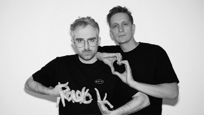 Steffen Israel und Felix Brummer von Kraftklub vor einer weißen Wand. Mit seinen Fingern formt Steffen "Radio K" für ihre Fritz-Radiosendung "Radio mit K". (Foto: Philipp Gladsome)